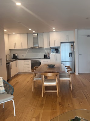 A modern kitchen with white cabinets, a wooden dining table, and a built-in microwave. - Waterfront escape with lake views, private pontoon & balcony, 5 mins to beach. (Tennyson)