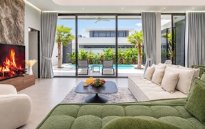 Una sala de estar moderna con un televisor de pantalla plana, un sofá verde, una mesa de centro con frutas y bebidas, y vistas a una piscina y zonas verdes al exterior.