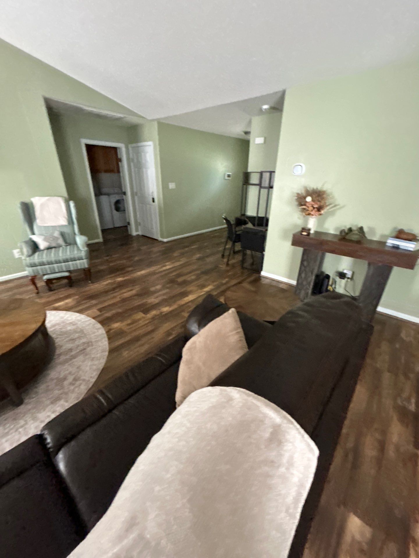 A living room with a dark brown sofa, a round coffee table, and a light-colored armchair. There is a dining area in the background with a table and chairs.