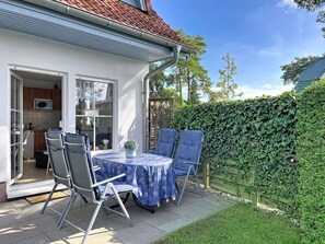 Eine Terrasse mit Tisch und Stühlen, eine durch eine offene Tür erkennbare Küche und ein grüner Heckenstreifen.