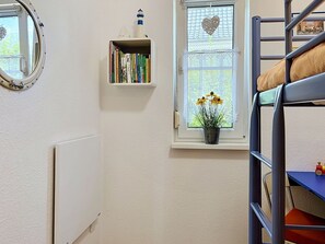 Ein Zimmer mit einem Etagenbett, einem Bücherregal, einem Heizkörper, einem Fenster mit Herzdeko und Blick ins Freie.