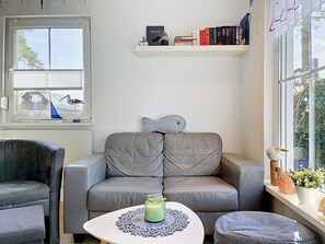 Sala de estar com sofá cinza, mesa redonda pequena com um jogo de toalhas decorativo, estante com livros e janela com vista para o verde.