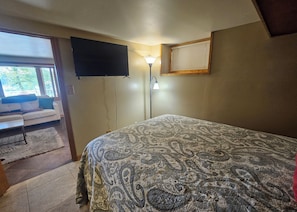 A bedroom with a bed, a TV mounted on the wall, a bedside lamp, and a view of the outdoors through a window. - Okauchee Lakefront Escape with Amazing Sunsets, Fire Pits, Deck & Pet Friendly (Nashotah)