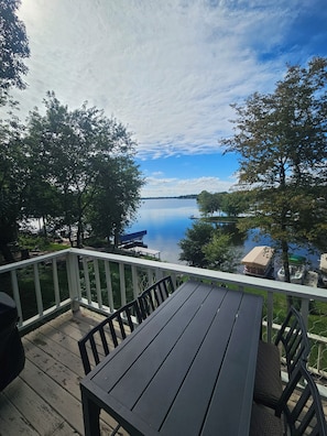 A wooden deck with a table and chairs, overlooking a calm lake with boats and trees. - Okauchee Lakefront Escape with Amazing Sunsets, Fire Pits, Deck & Pet Friendly (Nashotah)