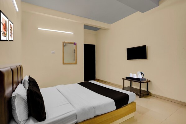A hotel room with a bed, a bedside table with a kettle, a flat-screen TV, and framed artwork on the wall.