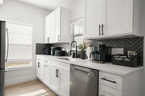 A modern kitchen with white cabinets, a stainless steel dishwasher, a coffee station, and a herringbone-patterned backsplash. - Private 3BR 2.5BA Home | Sleeps 9 | Fire Pit, BBQ + Fenced Yard | 15 Min to ATX (Austin)