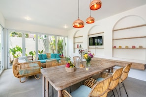 Un comedor con una mesa de madera, sillas de mimbre y un sofá. Hay plantas en macetas y un jarrón con flores sobre la mesa.