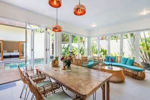 Amplia sala de estar con una gran mesa de comedor, mobiliario de mimbre y vistas a una piscina y follaje.