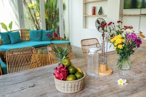 Un comedor con una mesa de madera, sillas de mimbre, una cesta de mimbre con frutas, una jarra de vidrio para agua y un florero con flores.