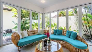 A room with a curved sofa set, a glass coffee table, and large windows offering a view of greenery outside.