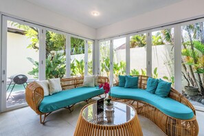 Una habitación con un sofá curvo, una mesa de centro de vidrio y amplios ventanales con vistas a zonas verdes.