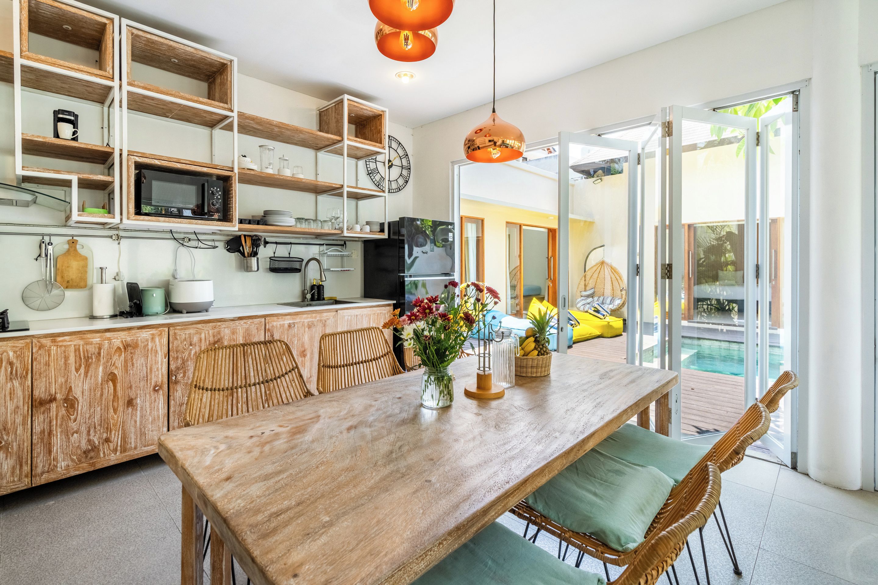 Een moderne keuken met houten kastjes, een eethoek met een houten tafel, en een zwembad zichtbaar door grote glazen deuren.