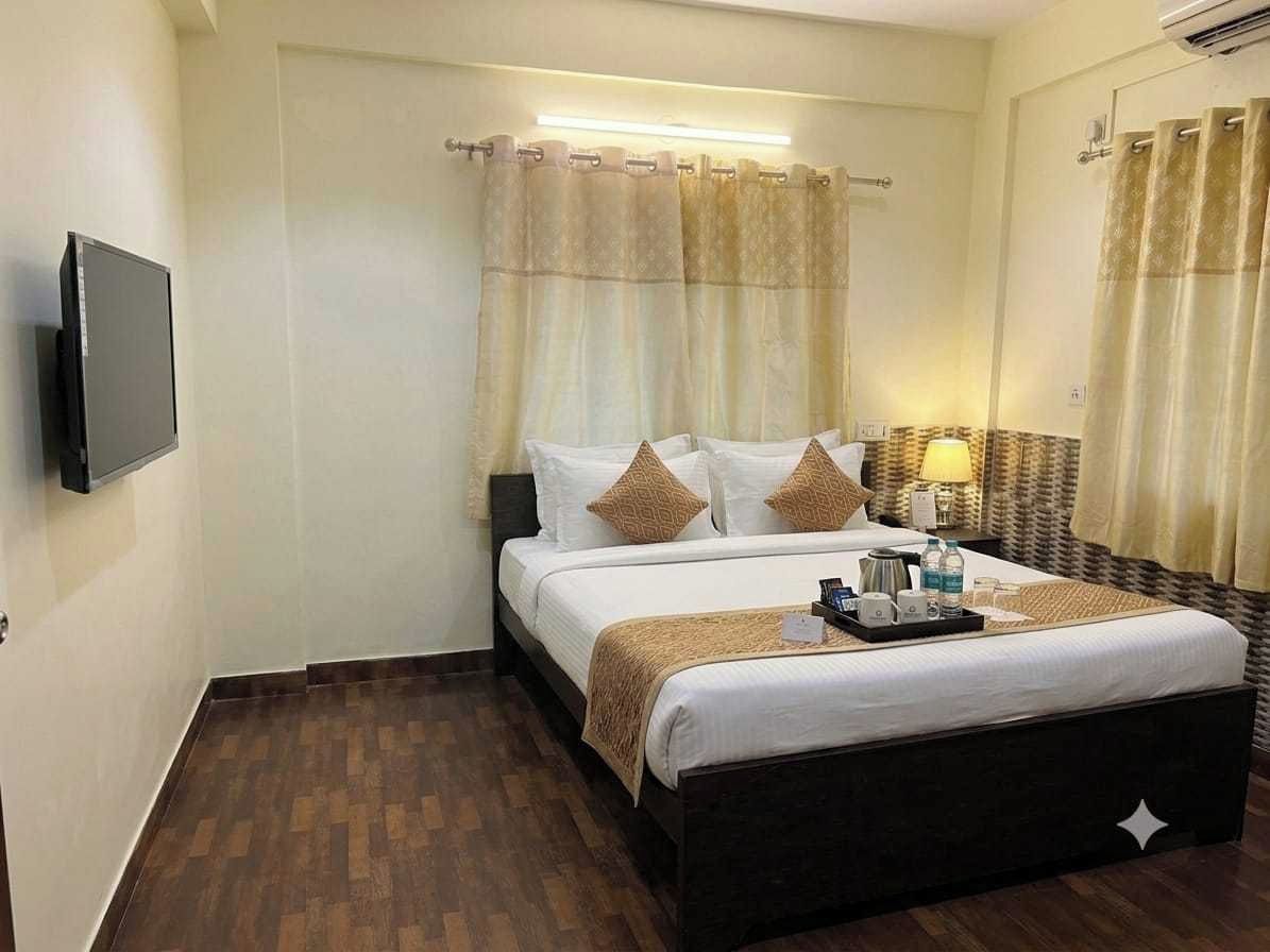 A hotel room with a bed, a flat-screen TV, a bedside table with a lamp, and a window with curtains.