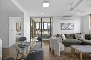 A modern living room with a glass dining table, a green sofa, and a city view through large windows. - Walk to King St & RBWH | Parking (Bowen Hills)