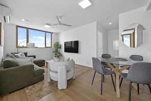 A modern living room with a green sofa, a white armchair, a dining table with chairs, a flat-screen TV, and a ceiling fan. - Walk to King St & RBWH | Parking (Bowen Hills)