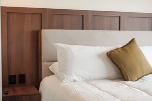 Una cama con ropa de cama blanca y almohada marrón, un cabecero de madera y una mesita de noche con un control remoto.