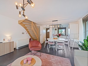 Una sala de estar moderna con una escalera de madera, un comedor con mobiliario blanco y una cocina visible a través de un puerta abierta.