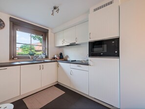 A modern kitchen with white cabinets, a black microwave, and a window with a view of greenery. - Modern holiday home with terrace by the coast (De Panne)