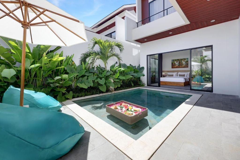 A modern outdoor pool area with a dining table set for two, surrounded by lush greenery and a large umbrella.