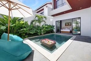 A modern outdoor pool area with a dining table set for two, surrounded by lush greenery and a large umbrella.