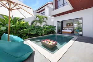 Un área de piscina moderna al aire libre con una mesa de comedor para dos, rodeada de exuberante vegetación y un amplio paraguas.