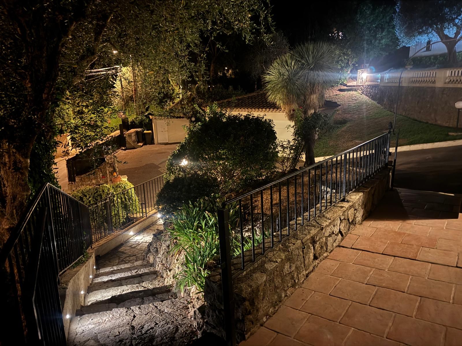 A stone staircase leading to a gated area with a metal railing, surrounded by greenery and a paved path.