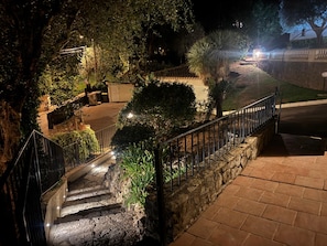 A stone staircase leading to a gated area with a metal railing, surrounded by greenery and a paved path. - Family home with a terrace in a quiet neighborhood in Pégomas (Pégomas)