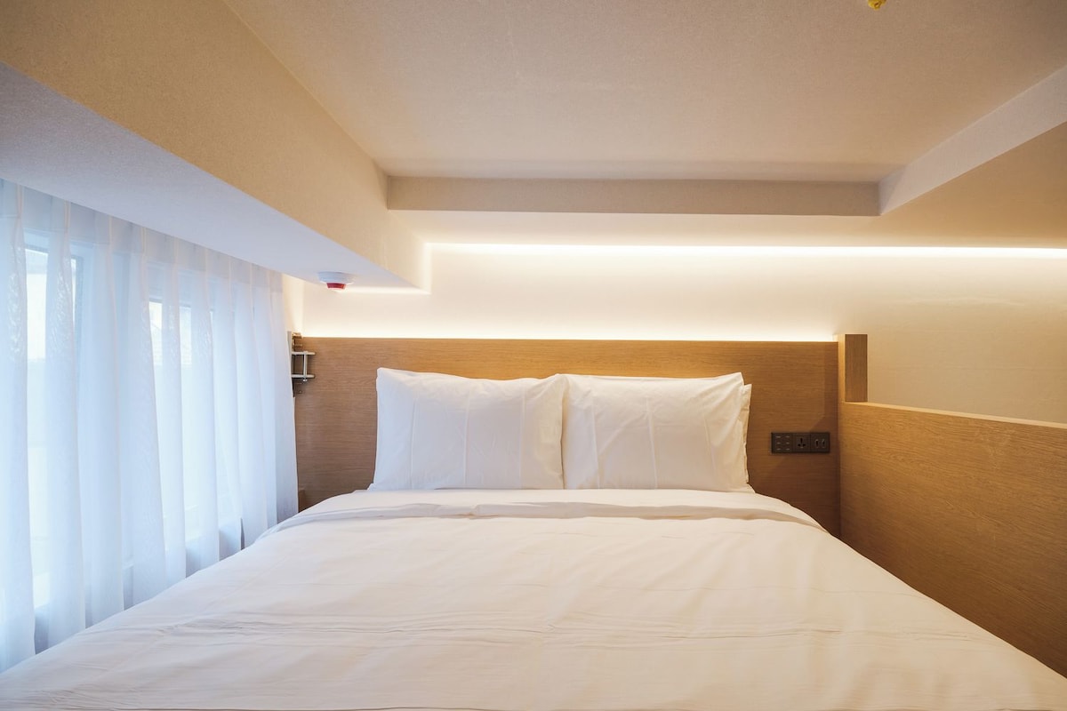 Habitación de hotel con cama, cabecera, mesita de noche y ventana con cortinas.