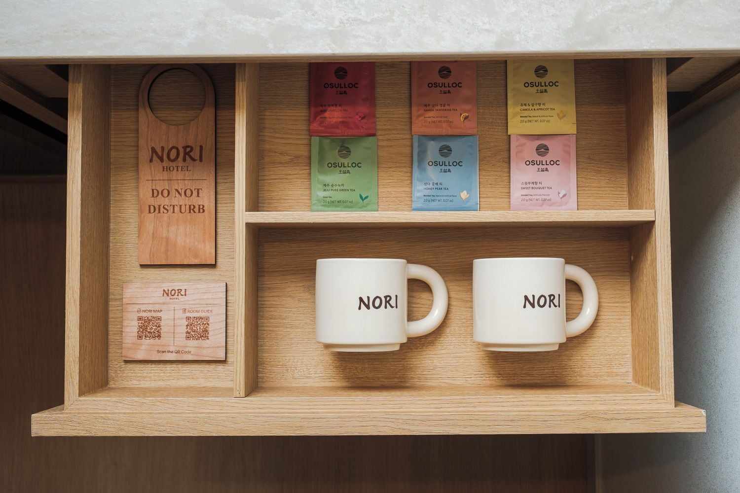 Una caja de madera con bolsitas de té y dos tazas blancas con la etiqueta 'NORI HOTEL', junto a un letrero de madera que dice 'NO MOLESTAR'.