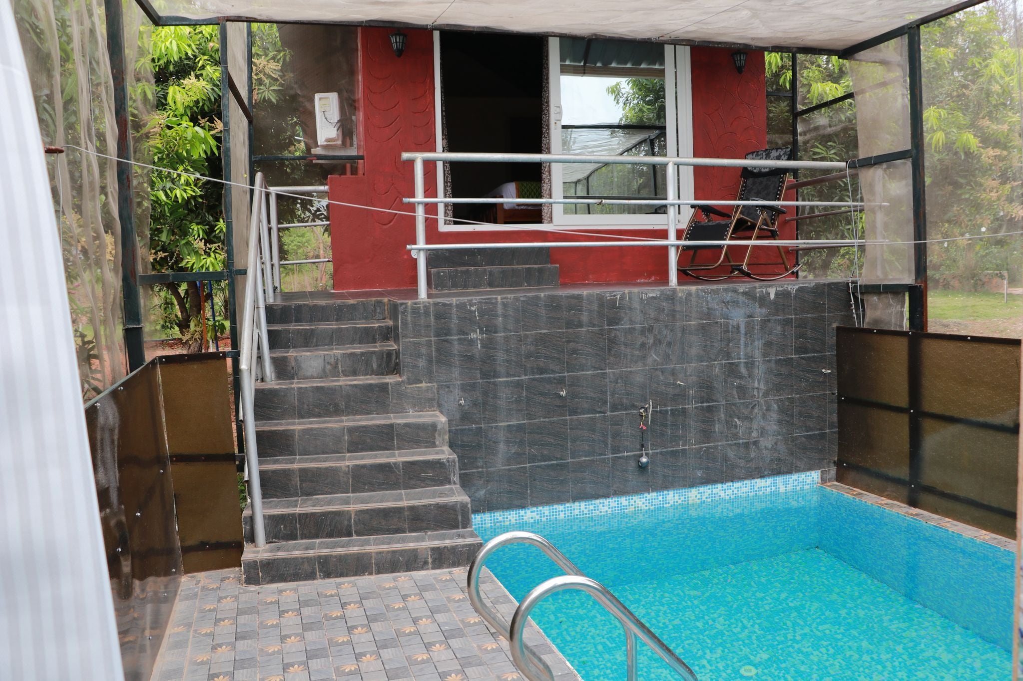 Una piscina con cerramiento de vidrio, escalones que conducen a un edificio rojo y una silla en el exterior.