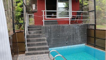 Una piscina con cerramiento de vidrio, escalones que conducen a un edificio rojo y una silla en el exterior.