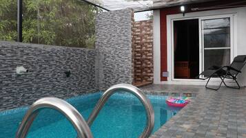 An outdoor swimming pool with metal pool ladders, a brick wall, and a red door leading to a covered area with two lounge chairs.