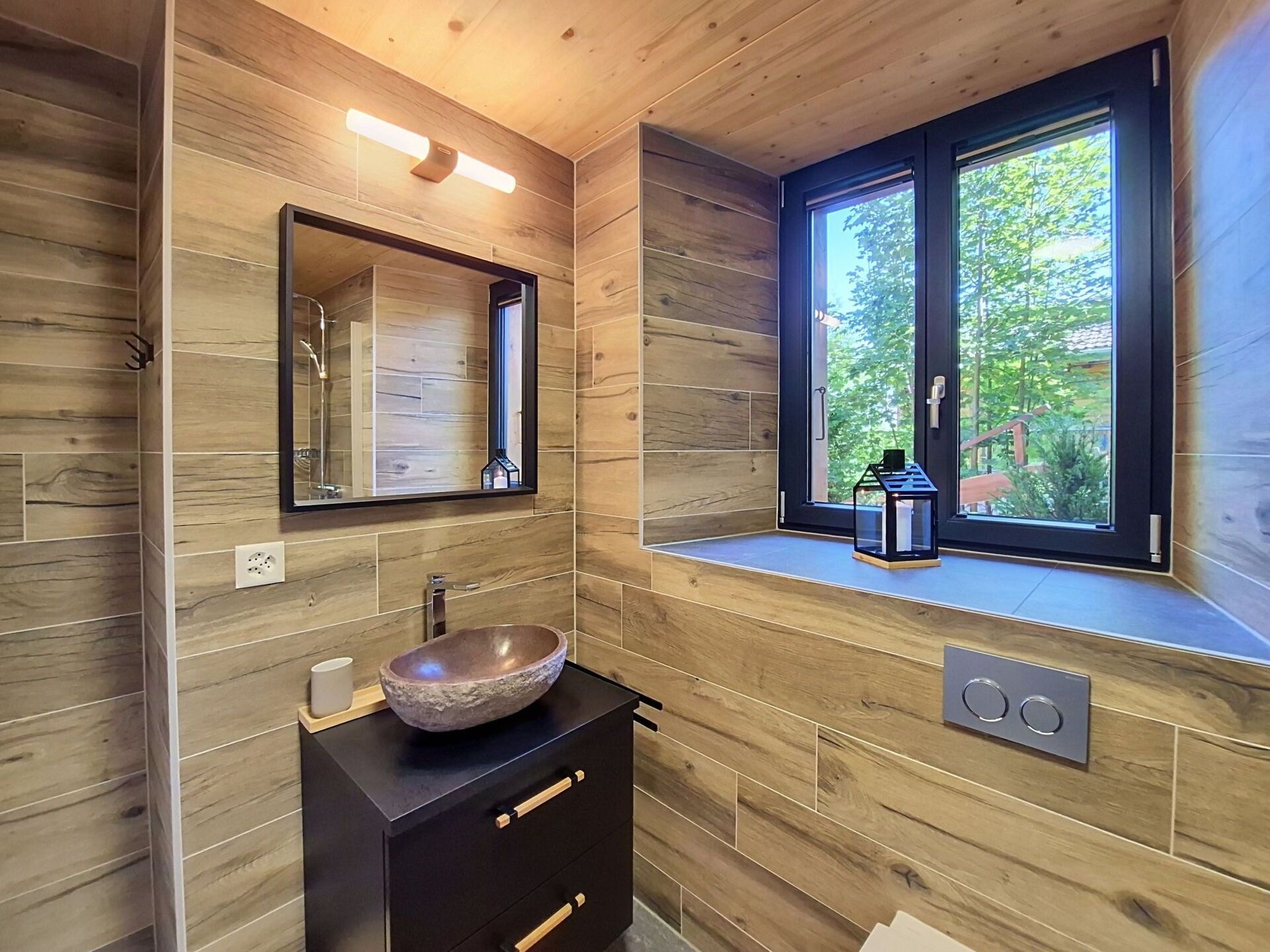 A modern bathroom with wooden walls, a large mirror, a vessel sink, and a window with a view of greenery.