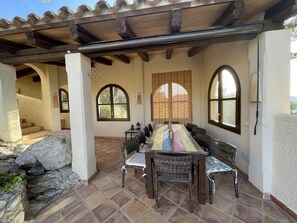 A covered patio with a long dining table and wicker chairs, large arched windows, and a stone wall. - "vista al Mar" Puerto Andratx (Andraitx)