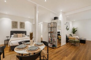 Une chambre d’hôtel moderne comprenant un lit, un coin repas avec une table dressée pour deux personnes, une bibliothèque et un coin salon avec un canapé et un fauteuil.