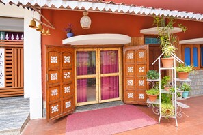 Une porte en bois traditionnelle ornée de panneaux décoratifs, un paillasson rouge à l’entrée et un présentoir métallique pour plantes, sur lequel sont disposées des plantes en pot.