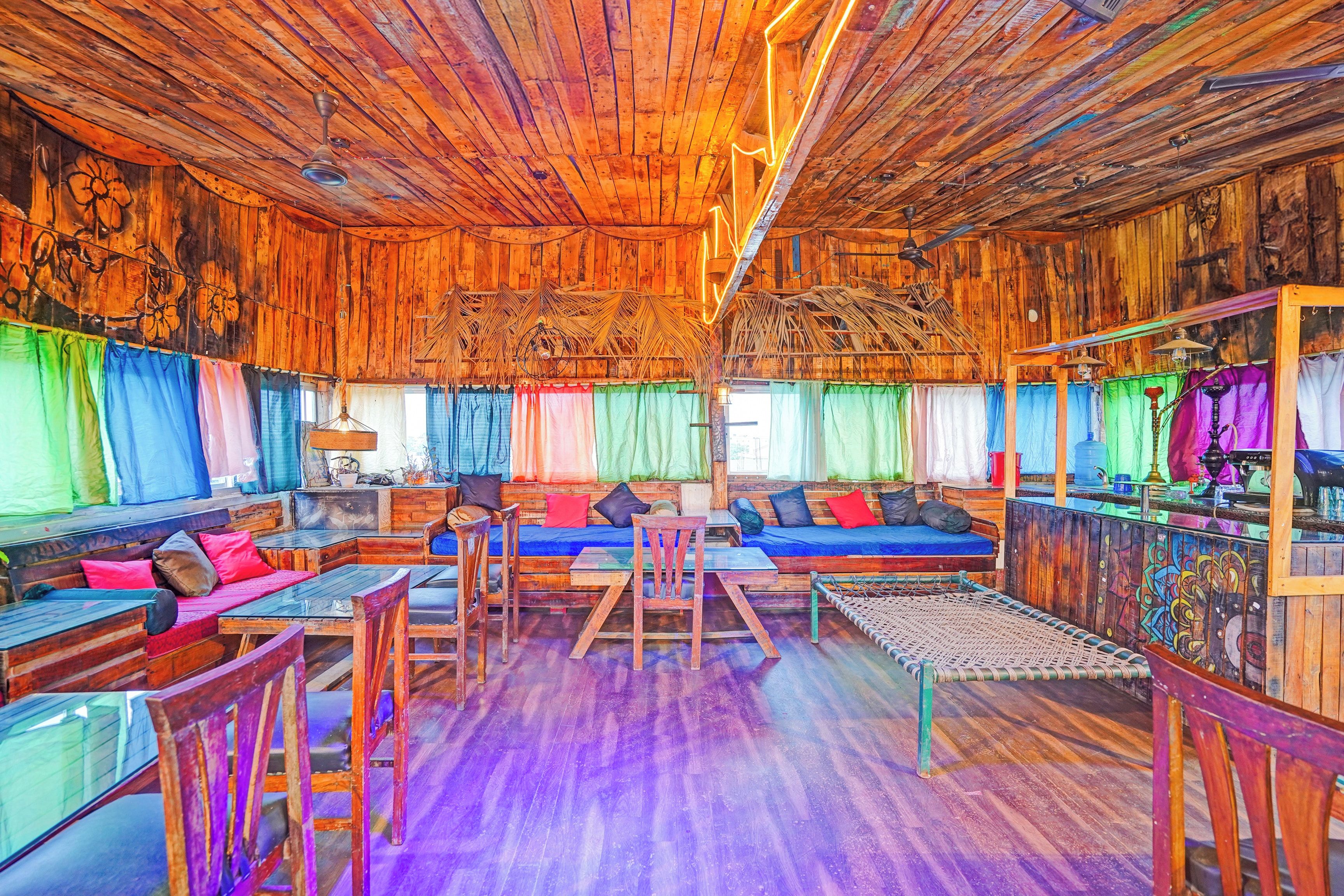 Un interior rústico con paredes y techos de madera, una zona de estar central con cojines, y un tratamiento de ventana colorido.