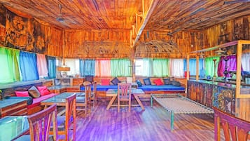 Un interior rústico con paredes y techos de madera, una zona de estar central con cojines, y un tratamiento de ventana colorido.
