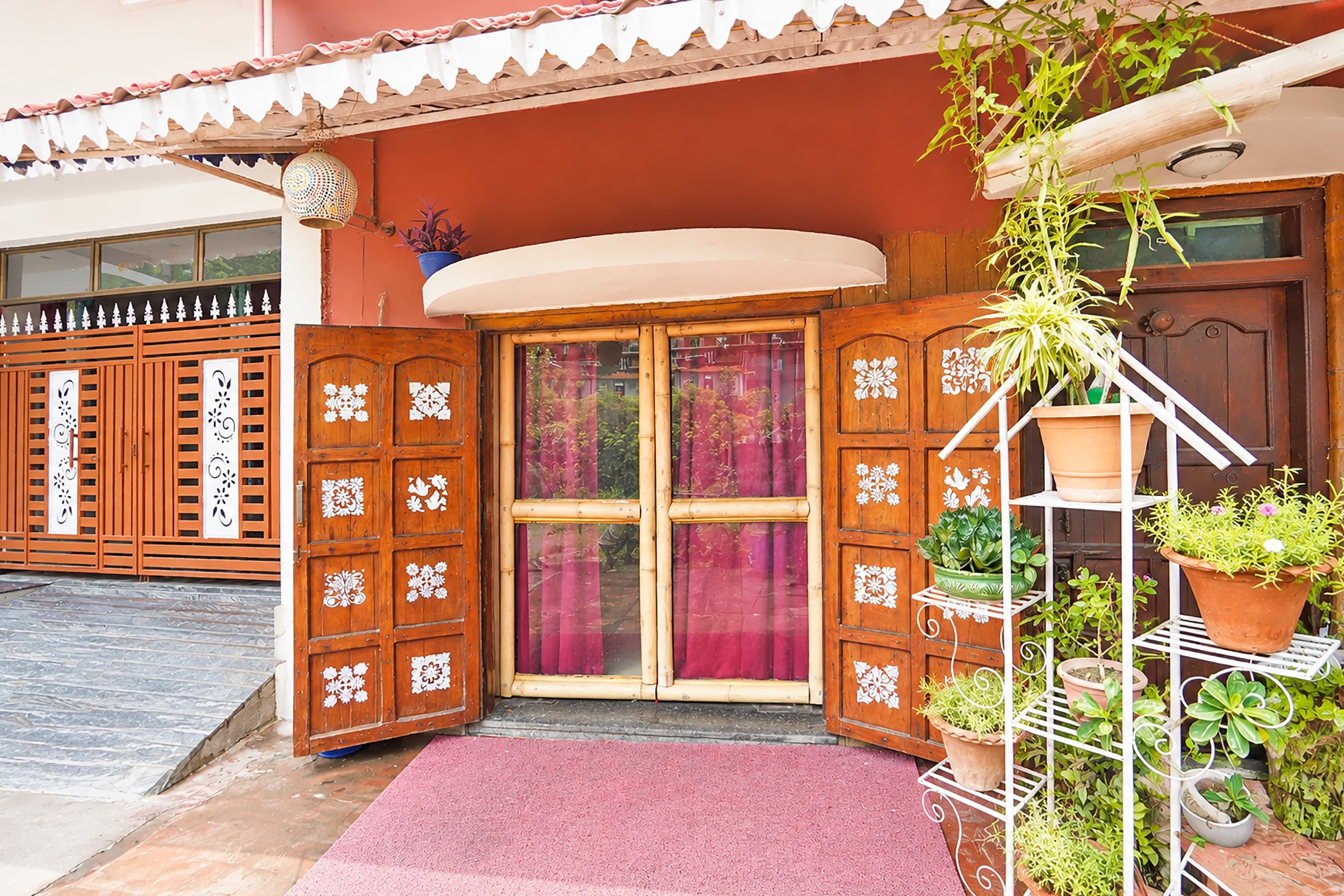 Una puerta de madera con elaborados diseños blancos, un felpudo rojo en la entrada y una planta en maceta sobre un soporte de metal blanco.