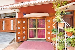 A wooden door with intricate white patterns, a red mat at the entrance, and a potted plant on a white metal stand.
