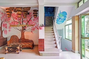A room with a large tree mural, a staircase with a blue mural, and a glass table with a woven base.
