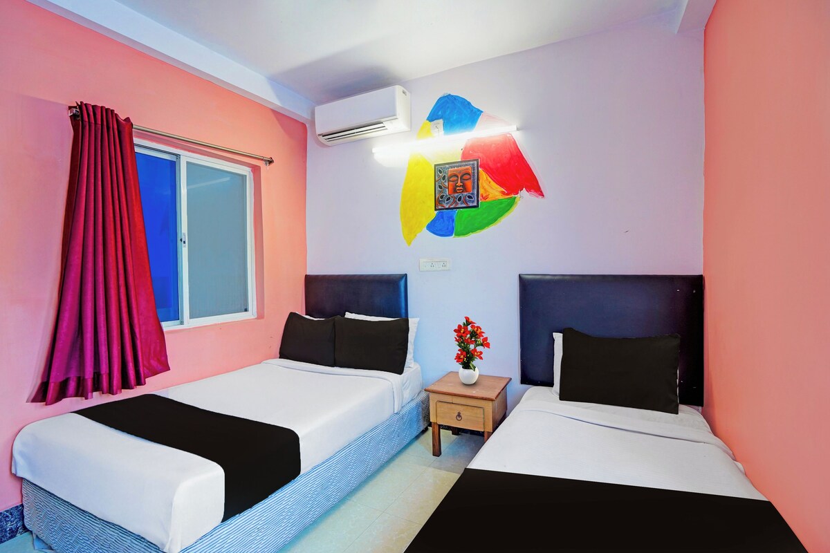 A hotel room with two beds, a window with curtains, an air conditioning unit, a wall-mounted artwork, and a small bedside table with a vase of flowers.