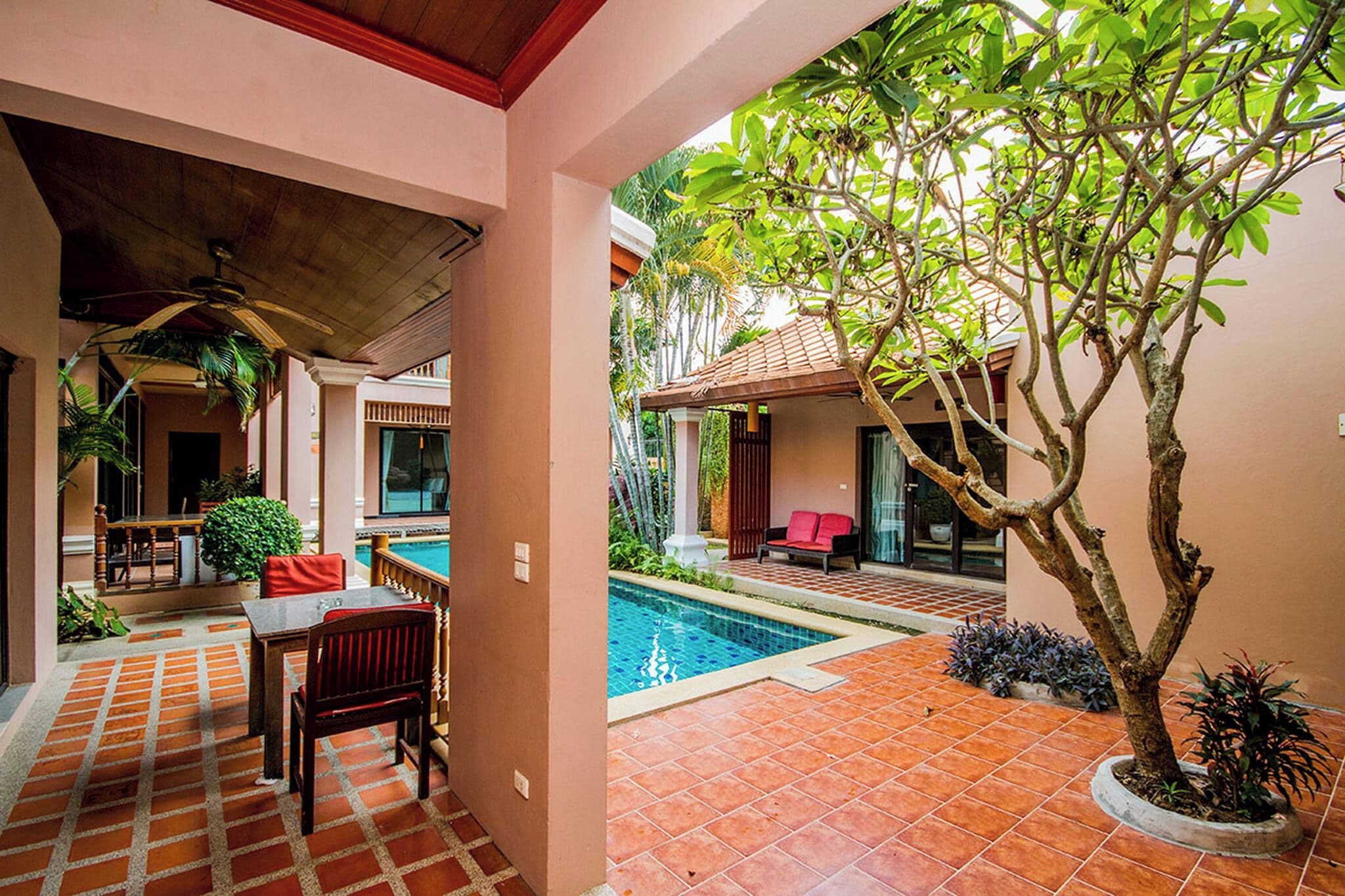 Zona de piscina con suelo de baldosas, una mesa con sillas y un árbol.
