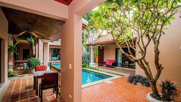Zona de piscina con suelo de baldosas, una mesa con sillas y un árbol.