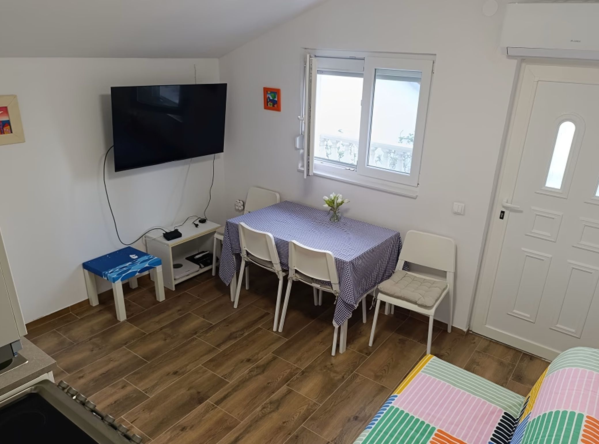 Una habitación con una televisión montada en la pared, una mesa de comedor con sillas, una ventana con vistas y una puerta.