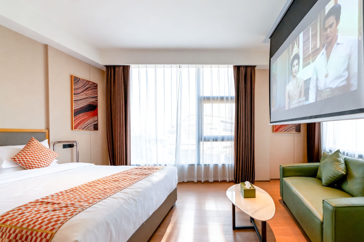 Una habitación de hotel con una cama, un sofá verde, una pantalla de televisión y una ventana con cortinas.