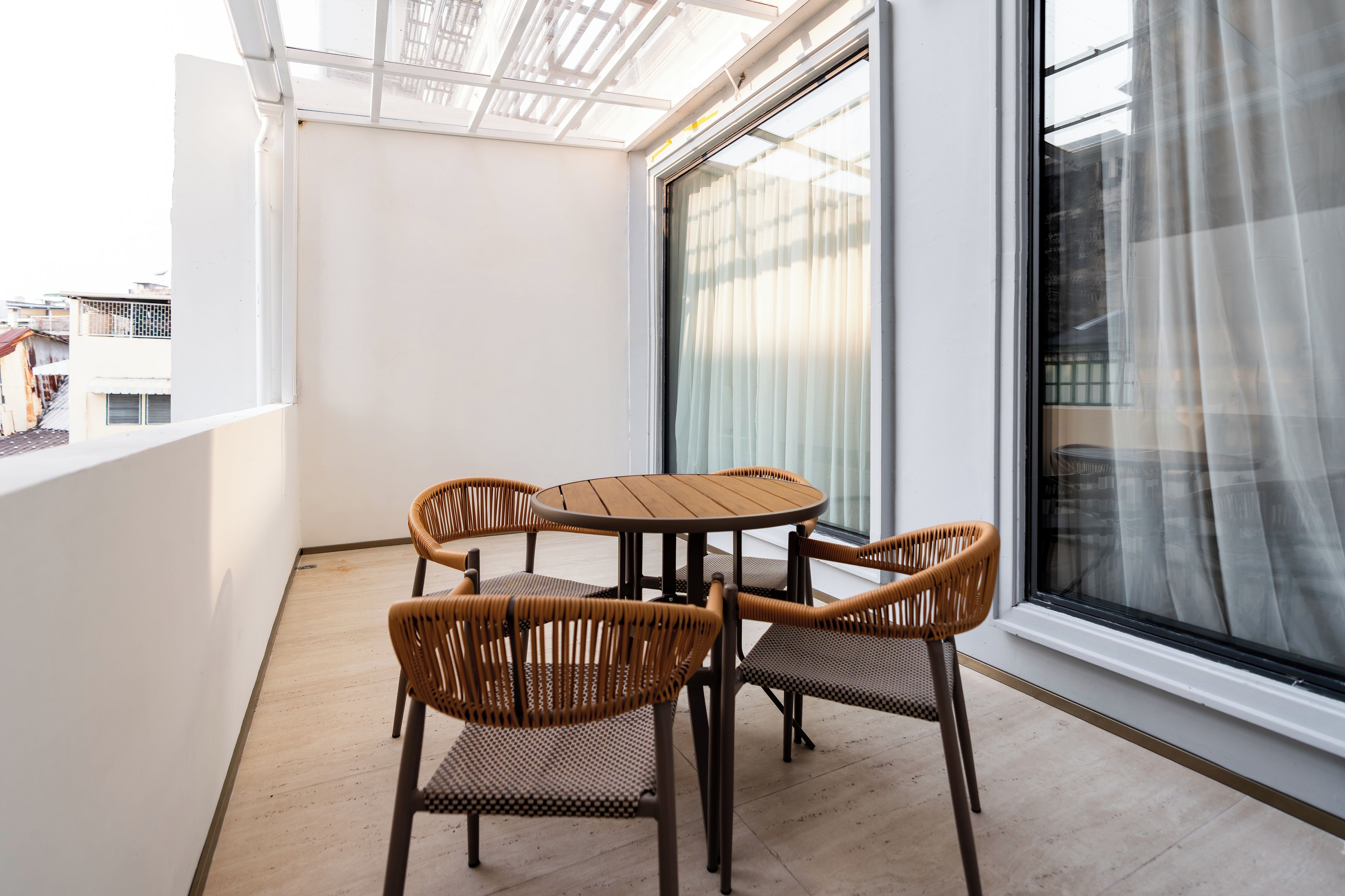 A balcony with a round table and four wicker chairs, a large window with sheer curtains, and a view of buildings.