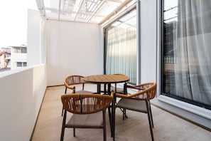 Un balcón con una mesa redonda y cuatro sillas de mimbre, un ventanal con cortinas translúcidas y vistas a edificios.