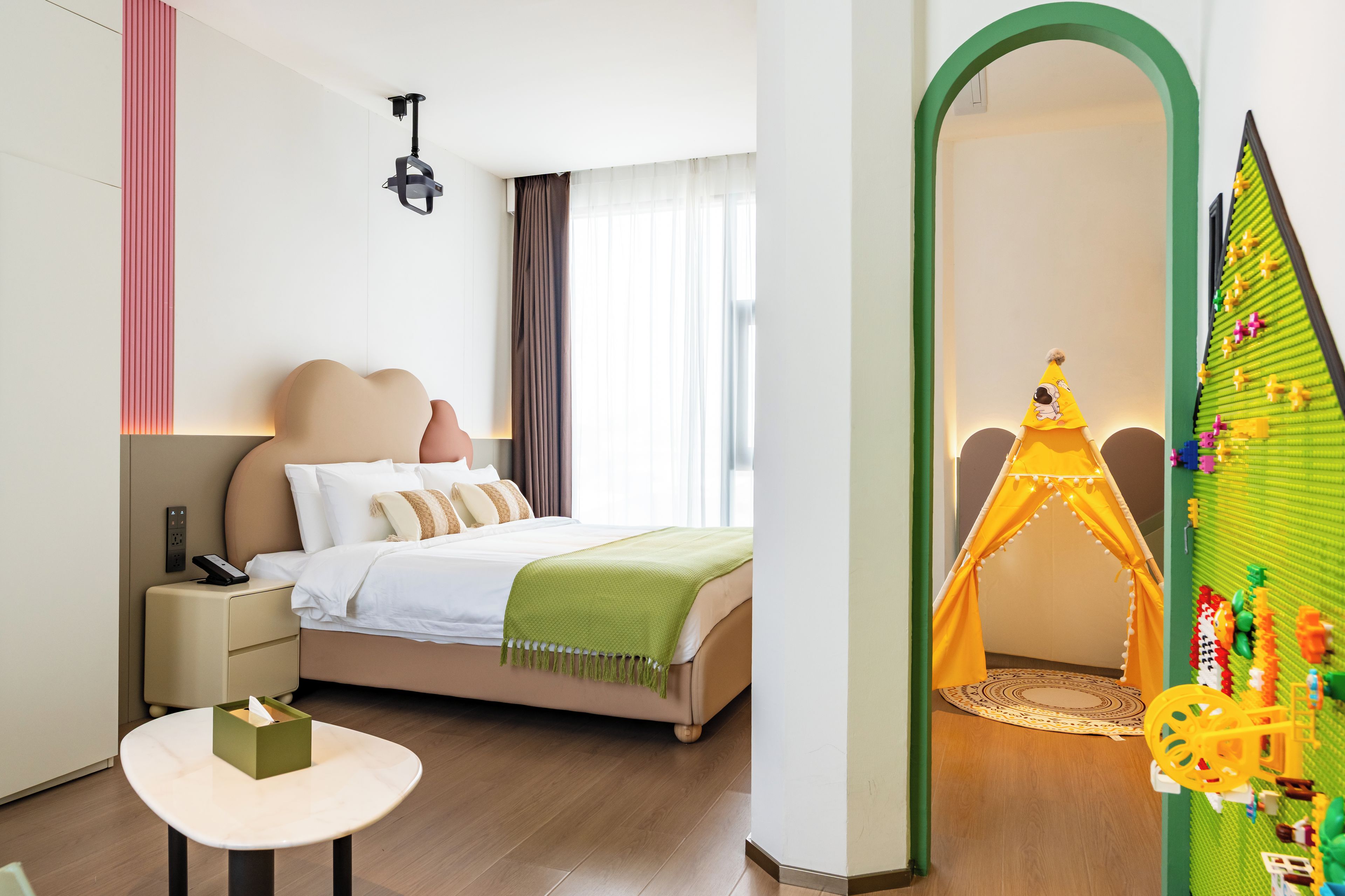 A modern hotel room with a bed, bedside table, and a green play tent in the corner.
