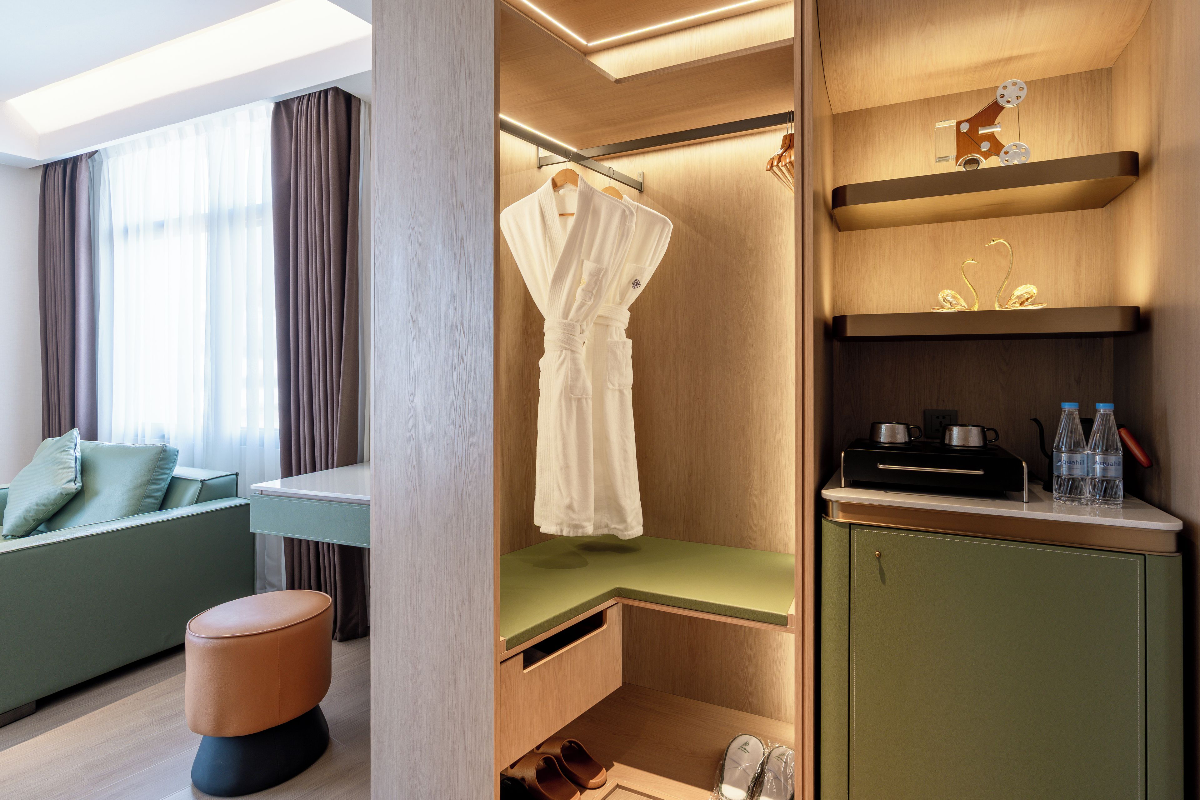 Ein modernes Hotelzimmer mit Sitzgelegenheit, einem aufgehängten Bademantel, einem Schminktisch mit Pflegeprodukten und einer Schuhablage.